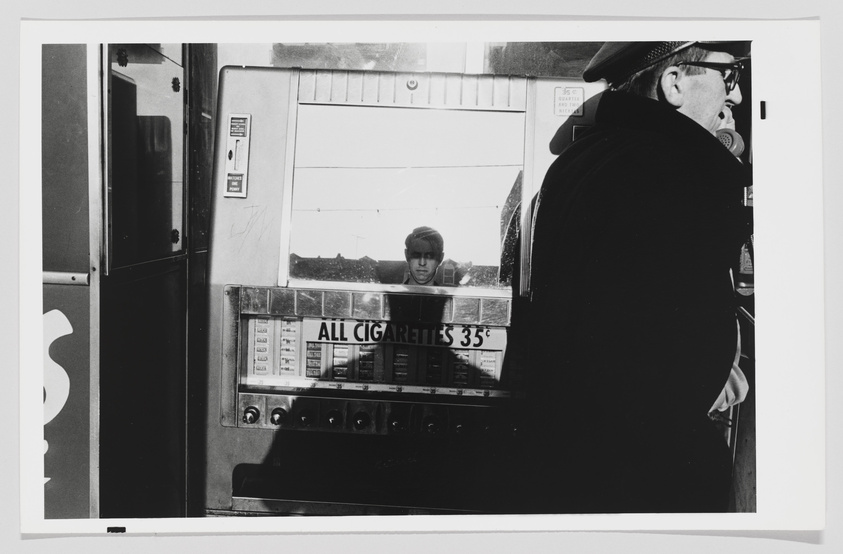 A black and white photograph capturing a moment at a cigarette vending machine. A man in a cap and glasses is seen from the side, possibly making a selection or purchase. Reflected in the glass of the machine is the face of a young person looking directly at the camera. The machine advertises "ALL CIGARETTES 35¢" with various brands and prices listed below. The setting appears to be indoors, with a glimpse of the outside visible in the reflection.