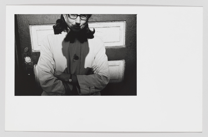 A black and white photograph of a person standing in front of a door, arms crossed, wearing glasses, a scarf, and a light-colored jacket. The person is looking directly at the camera with a slight smile. The door behind them has a dark color with a rectangular pattern.