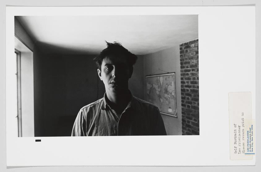 Black and white photograph of a young man standing in a room with a surprised expression, facing the camera. The room has a brick wall on the right and a framed map on the wall. The photo has a white border with a label on the bottom right indicating it is a self-portrait.