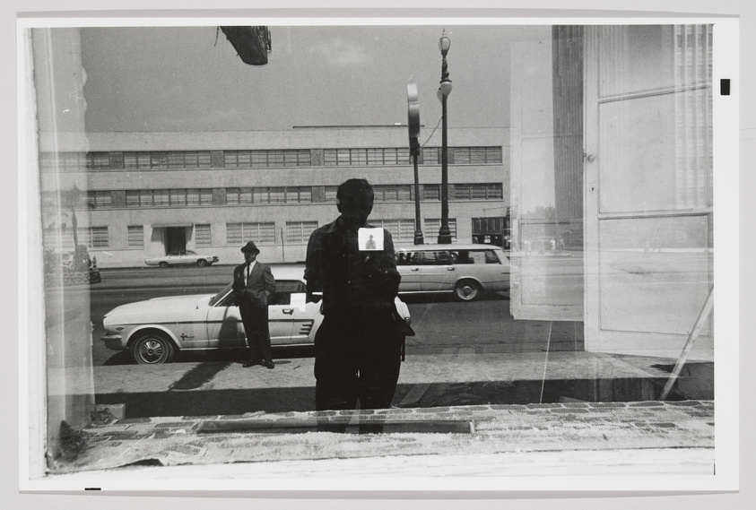 Black and white photograph capturing a reflection of a person holding a camera, seen through a glass pane with street scenes and buildings in the background. The silhouette of the photographer is centered, with a clear view of a man in a hat standing beside a classic car on the left and an industrial building across the street. The image has a layered appearance due to the reflective surface.