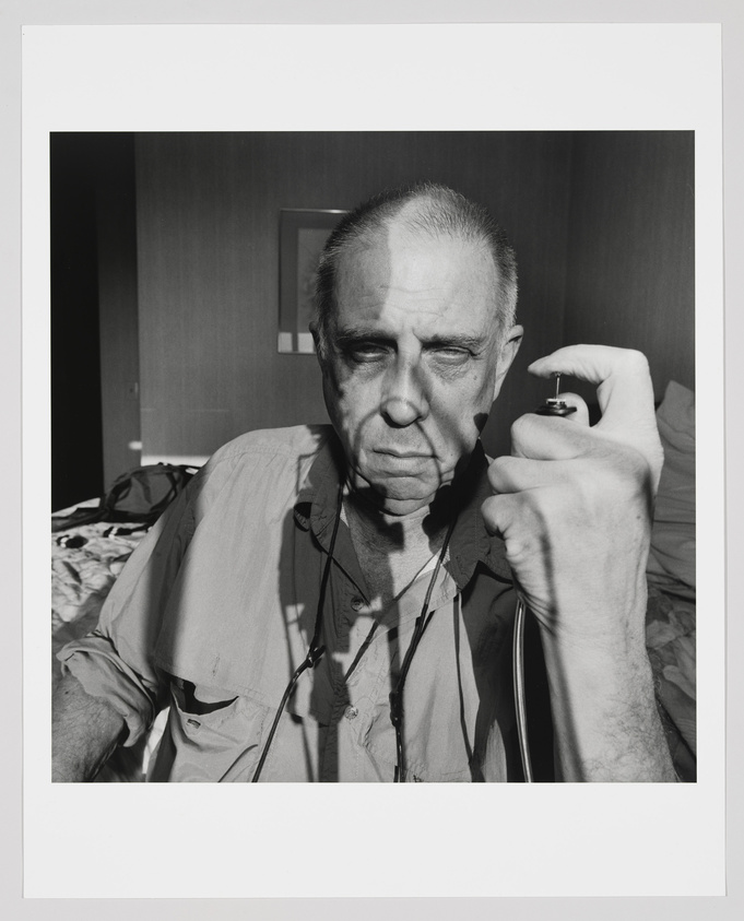 A black and white photograph of an older man with a stern expression, holding a camera's shutter release cable in his right hand. He is wearing a light-colored, short-sleeved shirt with a couple of unbuttoned buttons at the collar and has a lanyard around his neck. The background appears to be a hotel room with a bed and a mirror on the wall.