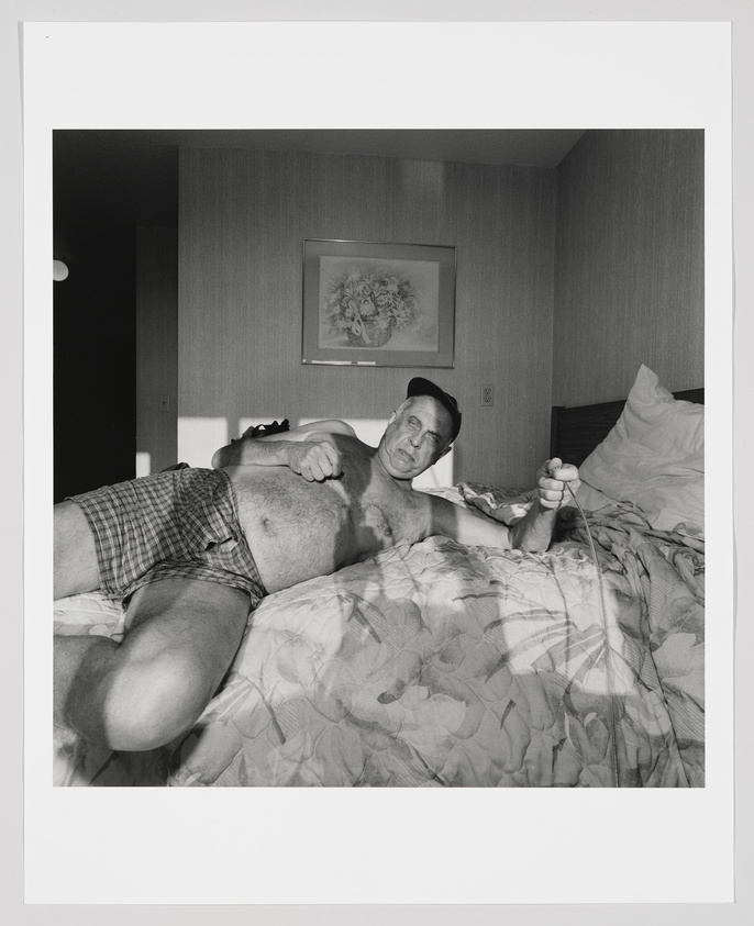 Black and white photograph of two men in a bedroom, one lying on his back on a bed with his shirt lifted to expose his belly, and the other sitting beside him, holding a stethoscope to the exposed belly. The room has a simple decor with a framed picture on the wall. Both men appear to be middle-aged, and the man with the stethoscope is looking at the camera with a serious expression.
