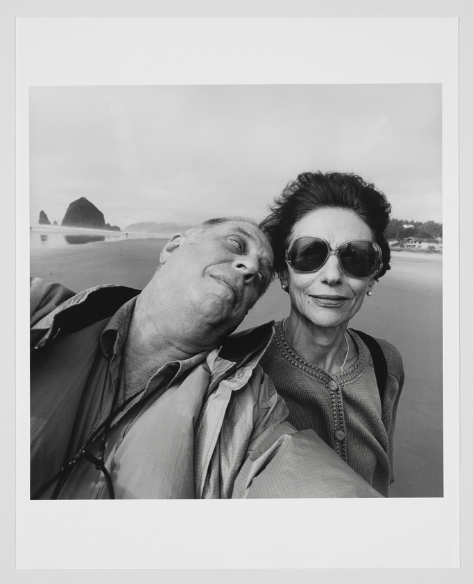 A black and white photo of an older couple taking a close-up selfie on a beach. The man is leaning his head towards the woman, eyes closed with a content expression, while the woman, wearing large sunglasses, looks directly at the camera with a slight smile. In the background, a tranquil beach scene with a distinctive rock formation in the water is visible under a cloudy sky.