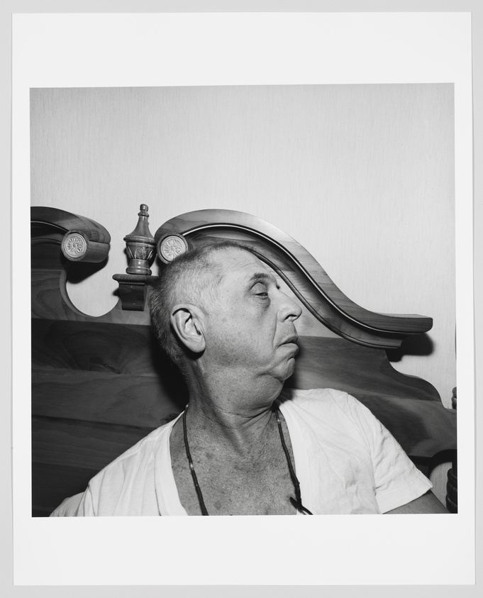 A black and white photograph of a man sitting in front of a wooden headboard, with his head turned to the side, revealing a coin stuck to his forehead. He is wearing a white undershirt and has a pensive expression on his face.