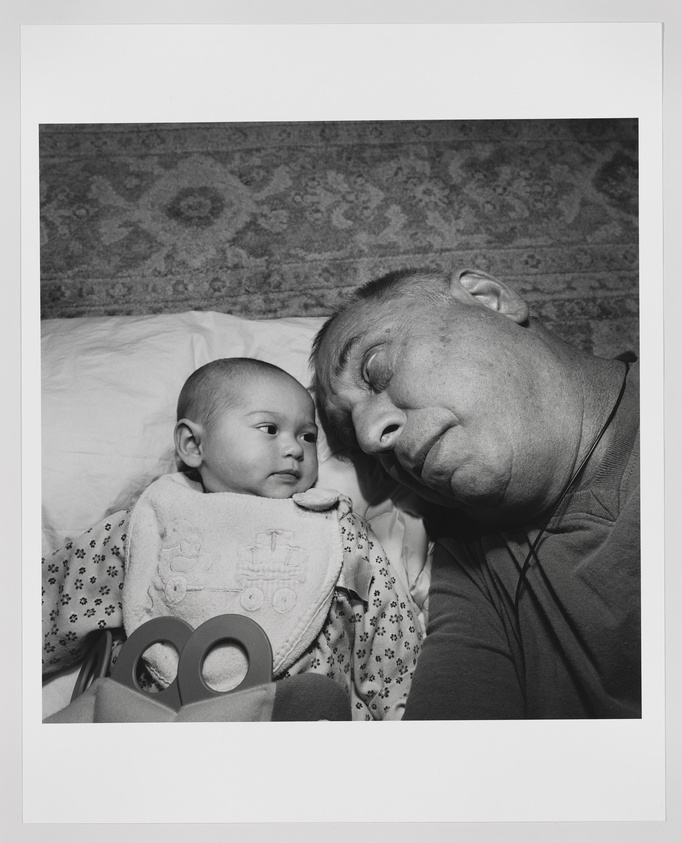 A black and white photograph capturing a tender moment between a baby and an adult, likely a family member. The baby, wearing a bib with a train design, looks calmly at the camera while the adult's face is turned towards the baby with a gentle, affectionate expression. Both are lying on a bed with a patterned carpet in the background.