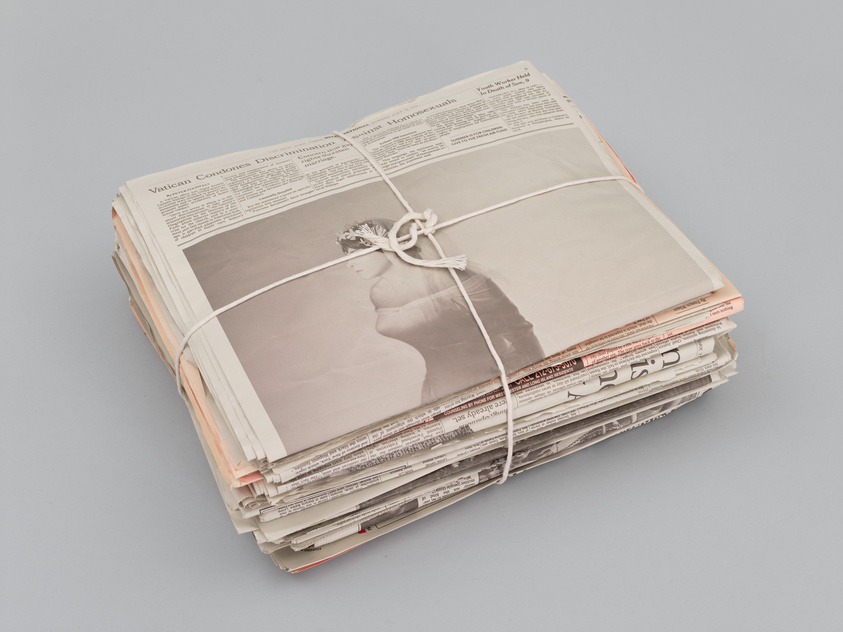 A stack of newspapers tied together with a white string, placed on a solid grey background. The visible top newspaper features an article with the headline "Vatican Condemns Discrimination Against Homosexuals."