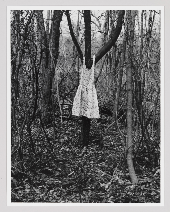 A black and white photograph depicting a leafless forest with a dress on a hanger suspended from a tree branch, creating the illusion of a headless figure standing among the trees. The dress is white with a floral pattern, and the surrounding forest floor is covered with fallen leaves.