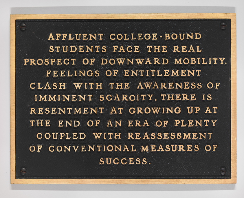 A black plaque with gold lettering that reads: "AFFLUENT COLLEGE-BOUND STUDENTS FACE THE REAL PROSPECT OF DOWNWARD MOBILITY. FEELINGS OF ENTITLEMENT CLASH WITH THE AWARENESS OF IMMINENT SCARCITY. THERE IS RESENTMENT AT GROWING UP AT THE END OF AN ERA OF PLENTY COUPLED WITH REASSESSMENT OF CONVENTIONAL MEASURES OF SUCCESS." The plaque is mounted on a wall with four screws, one in each corner.
