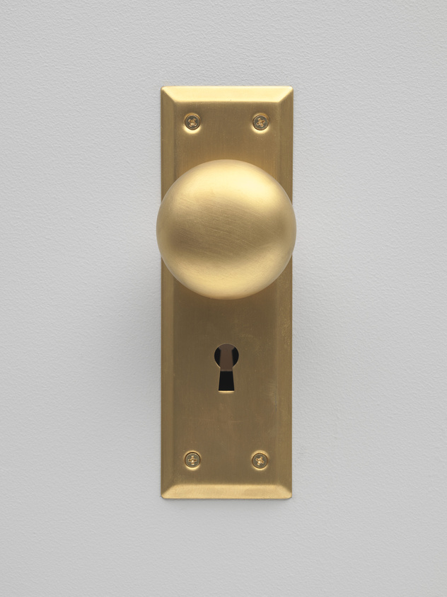 A brass door knob on a matching brass plate with a keyhole, mounted on a light gray wall.