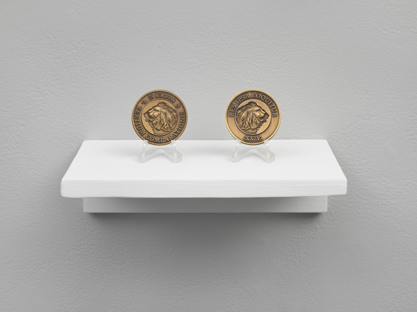 Two commemorative coins with embossed lion profiles are displayed on small clear plastic stands on a white shelf against a grey wall. The coin on the left has the inscription "IN MEMORY OF BRUTUM COMPANIONS," while the coin on the right reads "RESPERSA LOQUITUR XXIV."