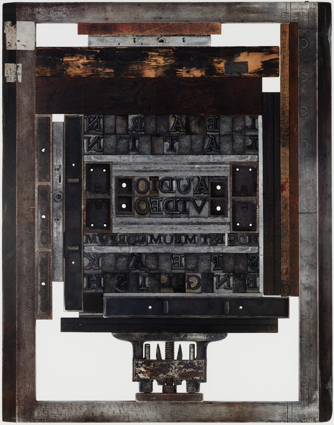 An image of a vintage printing press chase locked up with metal and wooden type blocks, some of which are reversed, showing letters and words in mirror image. The assembly is set against a white background, highlighting the textures and patina of the metal and wood components.