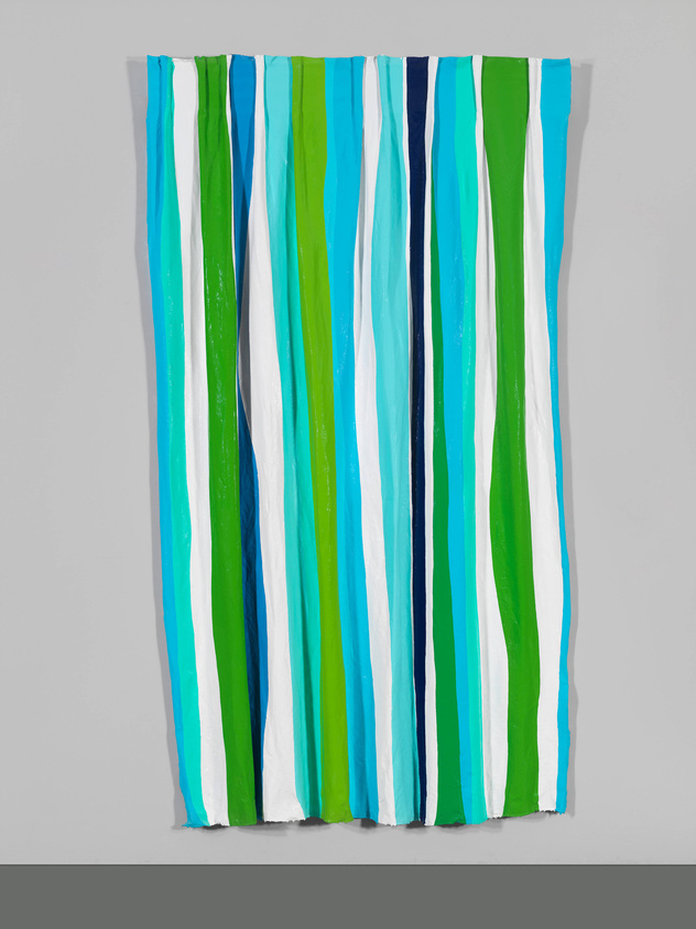 A modern art piece featuring vertical stripes in various shades of blue, green, and white, with a textured, three-dimensional appearance, hanging against a grey wall.