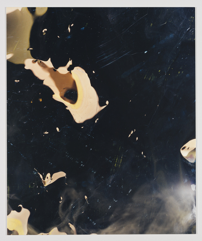 Large dark surface with scattered tan paint splatters and smudges, creating an abstract pattern. No text visible.