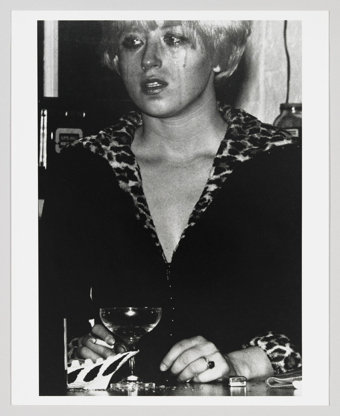 A black and white photograph of a person with short hair and dramatic makeup, including tears running down their cheeks. They are wearing a leopard print top with a deep neckline and are seated at a table with a cocktail glass in front of them. Their expression is somber and reflective.
