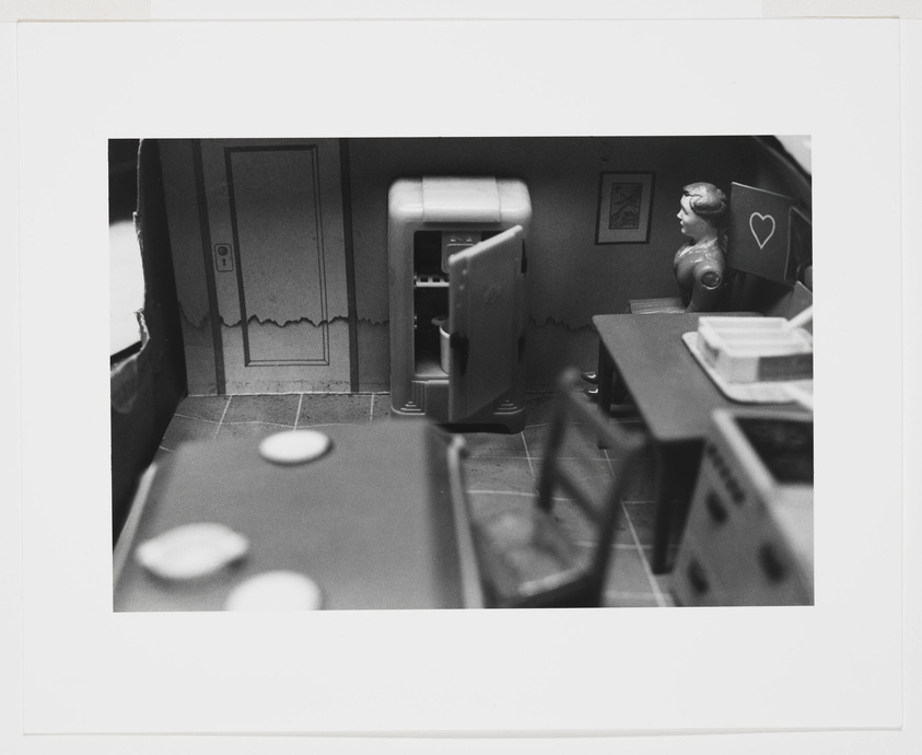 A black and white photo of a miniature kitchen scene with a vintage aesthetic, featuring a small fridge, a table with plates, and a figurine with a heart-shaped speech bubble next to it. The room includes details like a framed picture on the wall and a scalloped edge on the wall trim.