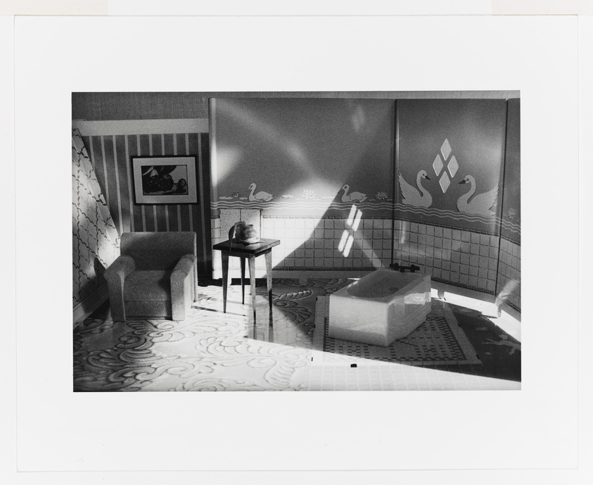 Black and white photo of a vintage bathroom with patterned tiles, a sunken tub, and a chair beside a small table.