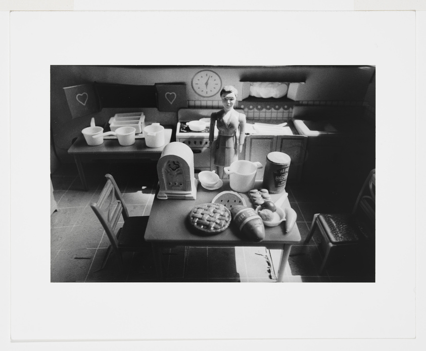 Black and white photo of a vintage toy kitchen with a doll and various miniature food items and utensils.