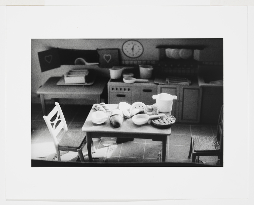 Monochrome photo of a miniature kitchen scene with a table set for a meal, a chair, and a clock on the wall.