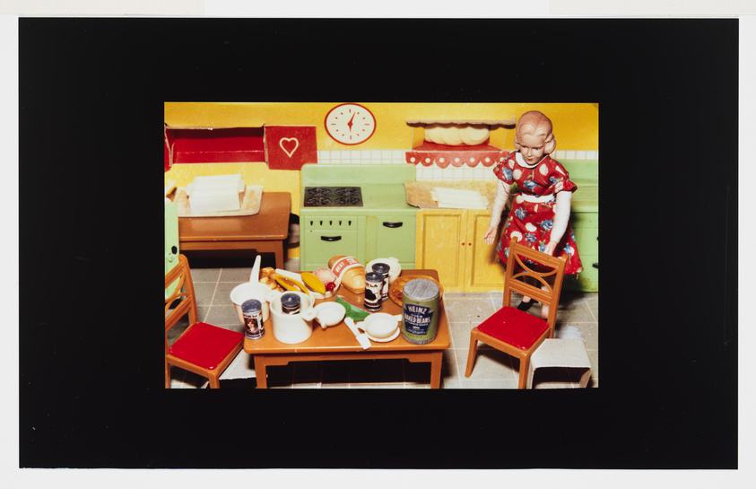 A doll in a colorful miniature kitchen with scattered food items and utensils on the table.