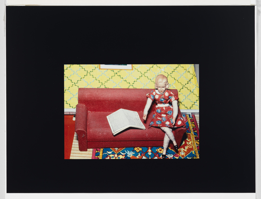 Doll in a red dress sits on a red couch with an open book, colorful rug below, and yellow patterned wallpaper behind.