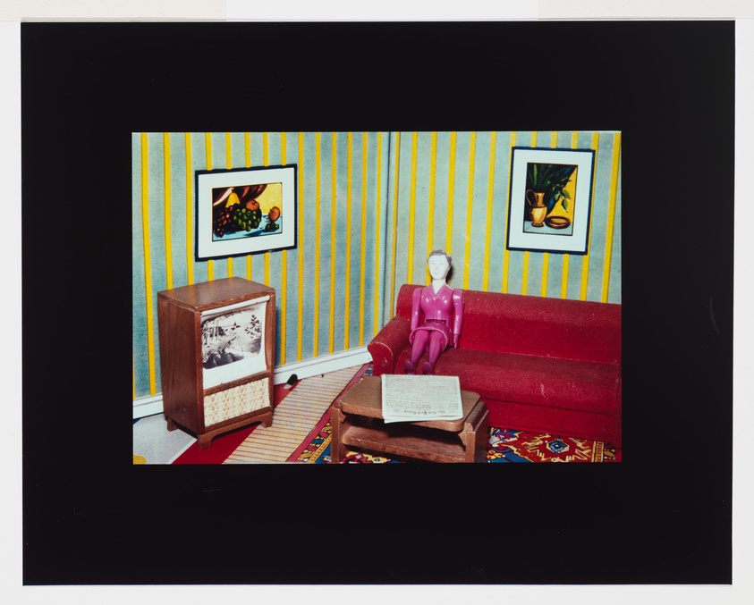 Vintage living room with striped wallpaper, red sofa, old TV, and a mannequin seated with a newspaper.