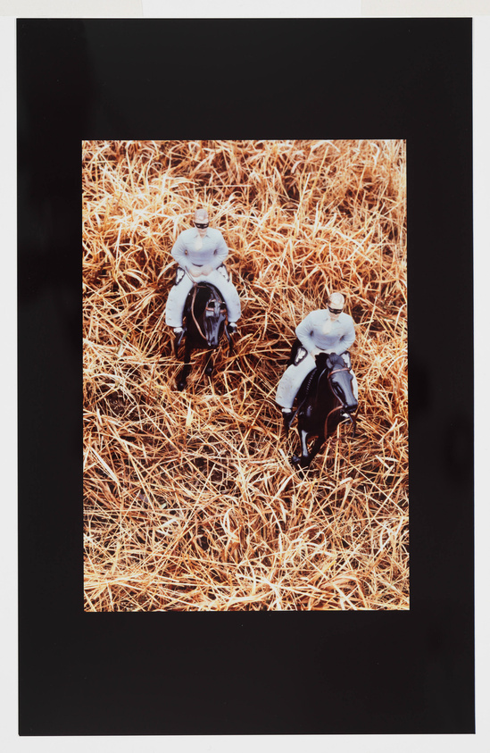 Two action figures resembling horseback riders are positioned on a bed of straw, with the top figure in a white outfit and the bottom figure in a black outfit, both on horseback, captured from an aerial perspective and framed with a black border.