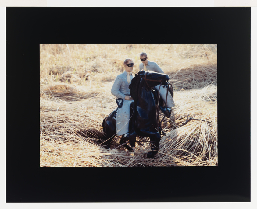 Two people in sunglasses posing with a black cloth on dry grass, framed by a black border.