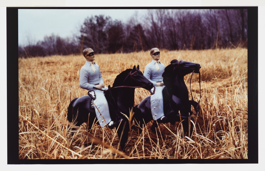 Two identical figures on horseback in a golden field, with a barren treeline in the background.