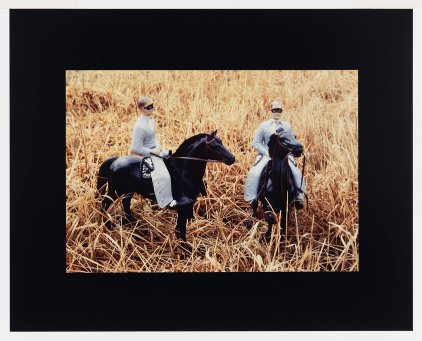 Two action figures resembling men in sunglasses and suits are mounted on toy horses in a field of dry, golden grass. The figures and horses are positioned side by side, facing the camera, evoking a surreal, miniature scene. The image is framed with a black border.