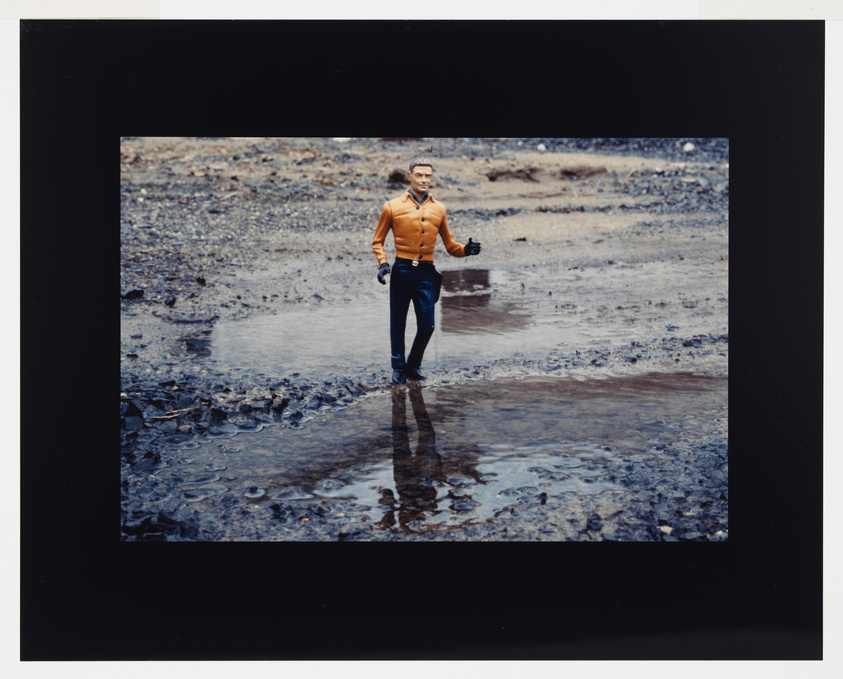 A toy figure in an orange shirt and black pants stands on a muddy, reflective surface, giving a thumbs-up gesture.