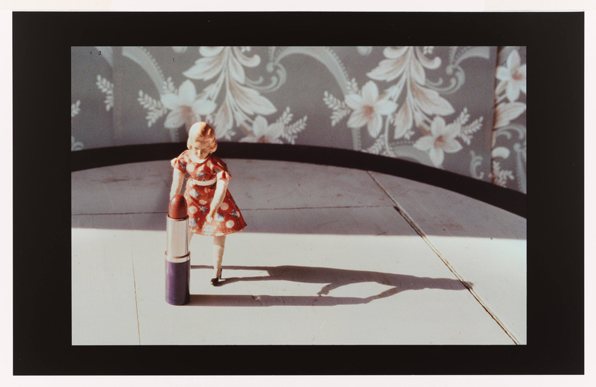 A vintage photograph featuring a small doll in a red and white dress standing next to an oversized lipstick on a stage with a floral backdrop, casting a long shadow on the surface.