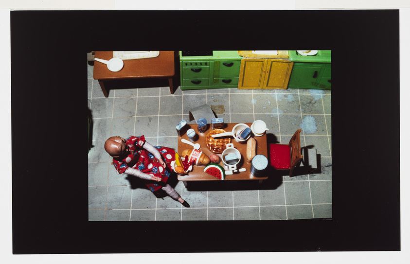 Bird's-eye view of a doll in a colorful dress sitting at a toy kitchen table with various miniature dishes and food items.
