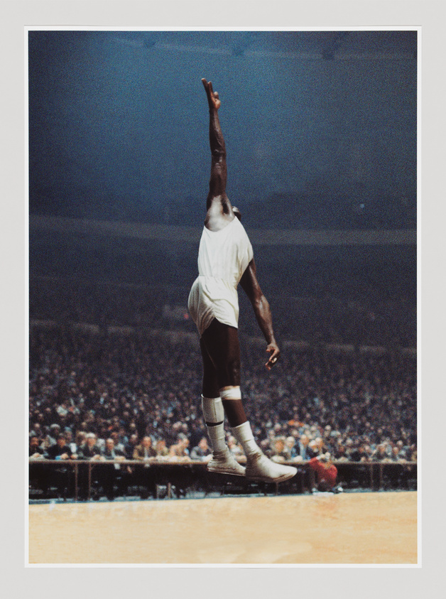 Basketball player in mid-air, reaching high with one arm extended, wearing a white uniform, with a packed stadium in the background.