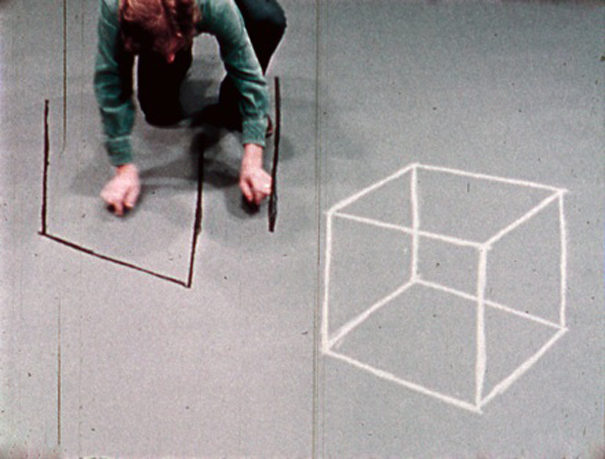 A person is crouched on the floor, drawing a square with chalk, next to a projected image of a three-dimensional cube on the wall.