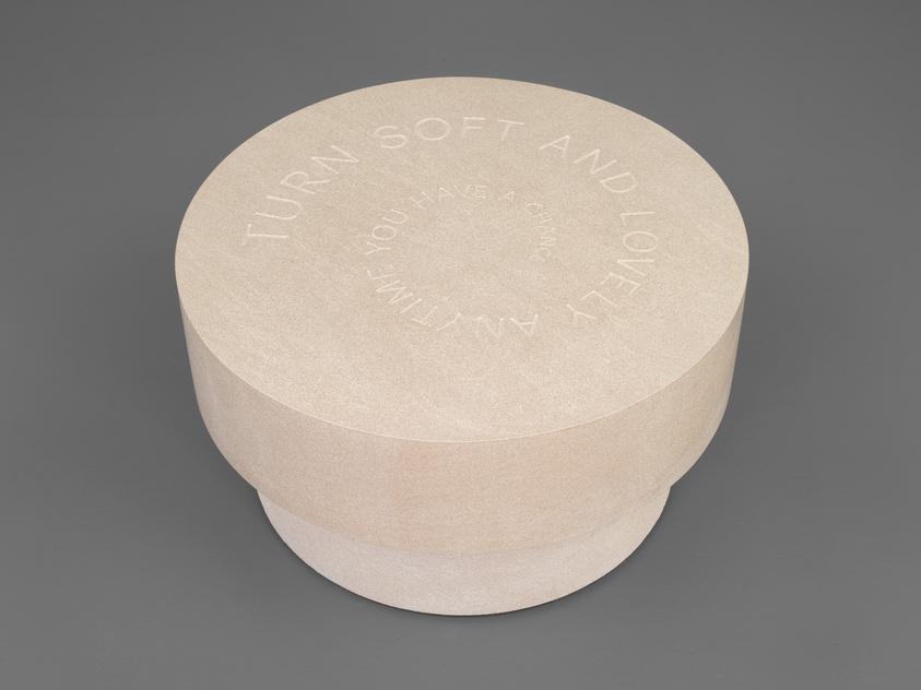 A round, beige-colored object with a flat top and curved sides sits on a gray surface. The top of the object has engraved text that reads "TURN SOFT AND LOVELY ANYTIME YOU HAVE A CHANCE."