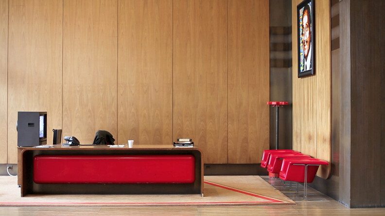 A modern reception area featuring a long red reception desk with a computer monitor, a black coat, and some papers on it. To the right, there are two red chairs against a wooden wall with a framed picture hanging above them. The floor is adorned with a large rug that has red accents matching the furniture.