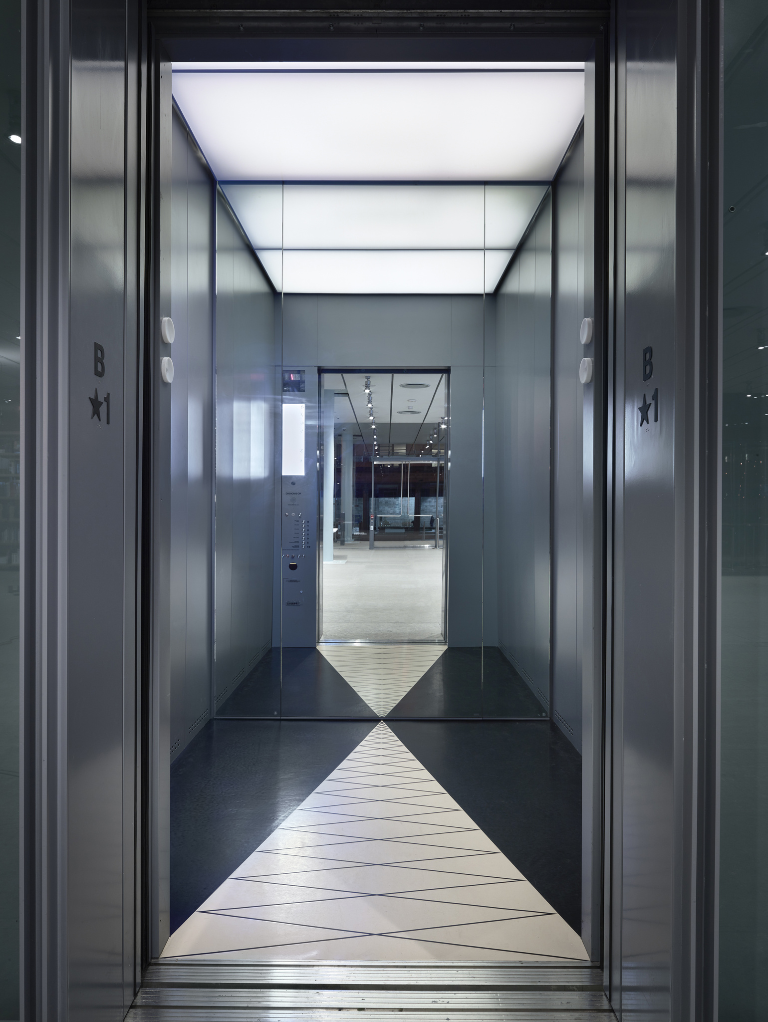 View from inside an elevator looking out into a modern building lobby with reflective floors and a geometric pattern on the threshold. The elevator has a lit ceiling panel and metallic interior with buttons and floor indicators visible on the side.