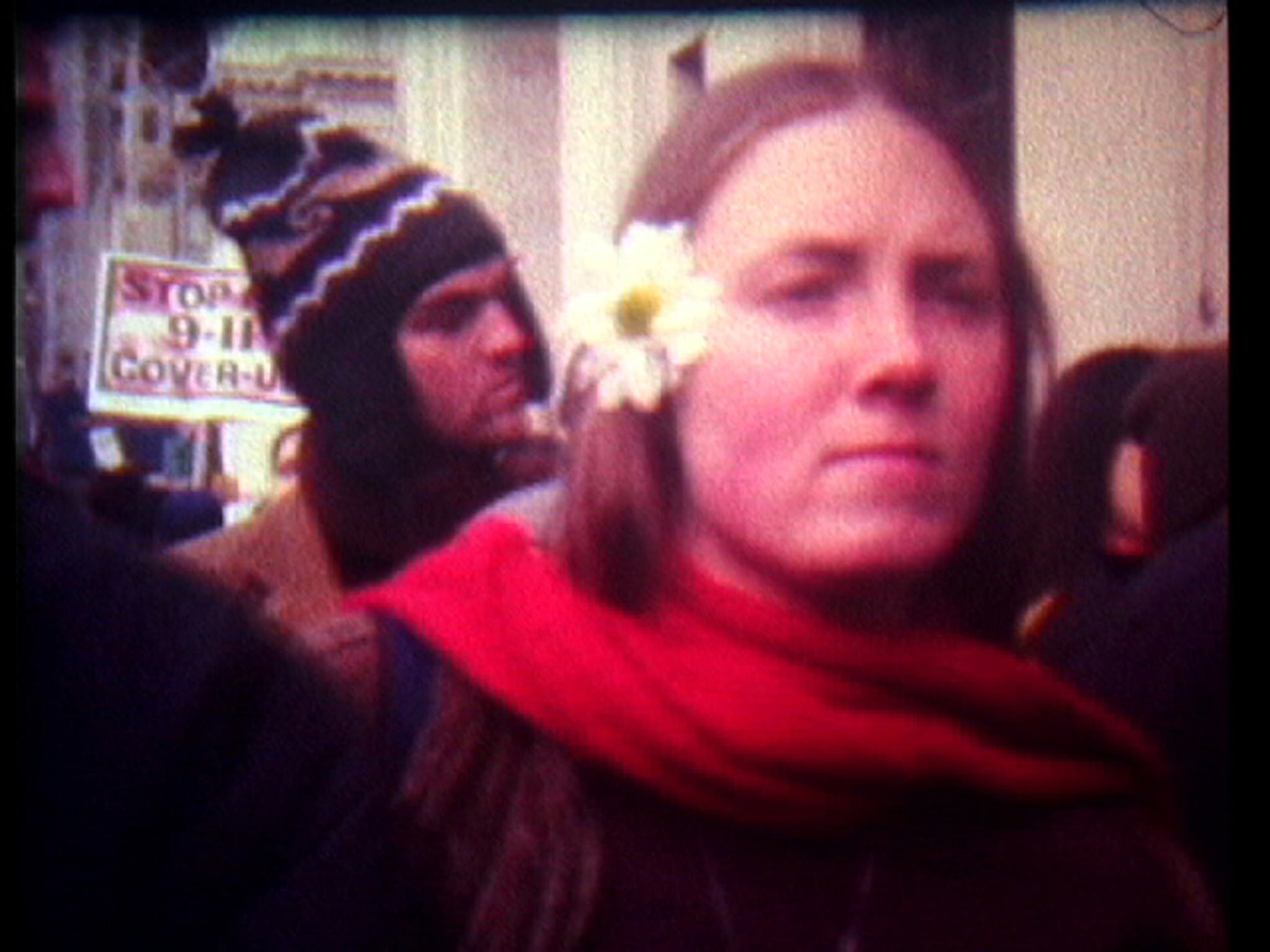 A grainy image of a woman with a solemn expression wearing a red scarf and a white flower in her hair. In the background, a person is holding a sign with the words "Stop 9-11 Cover-up."