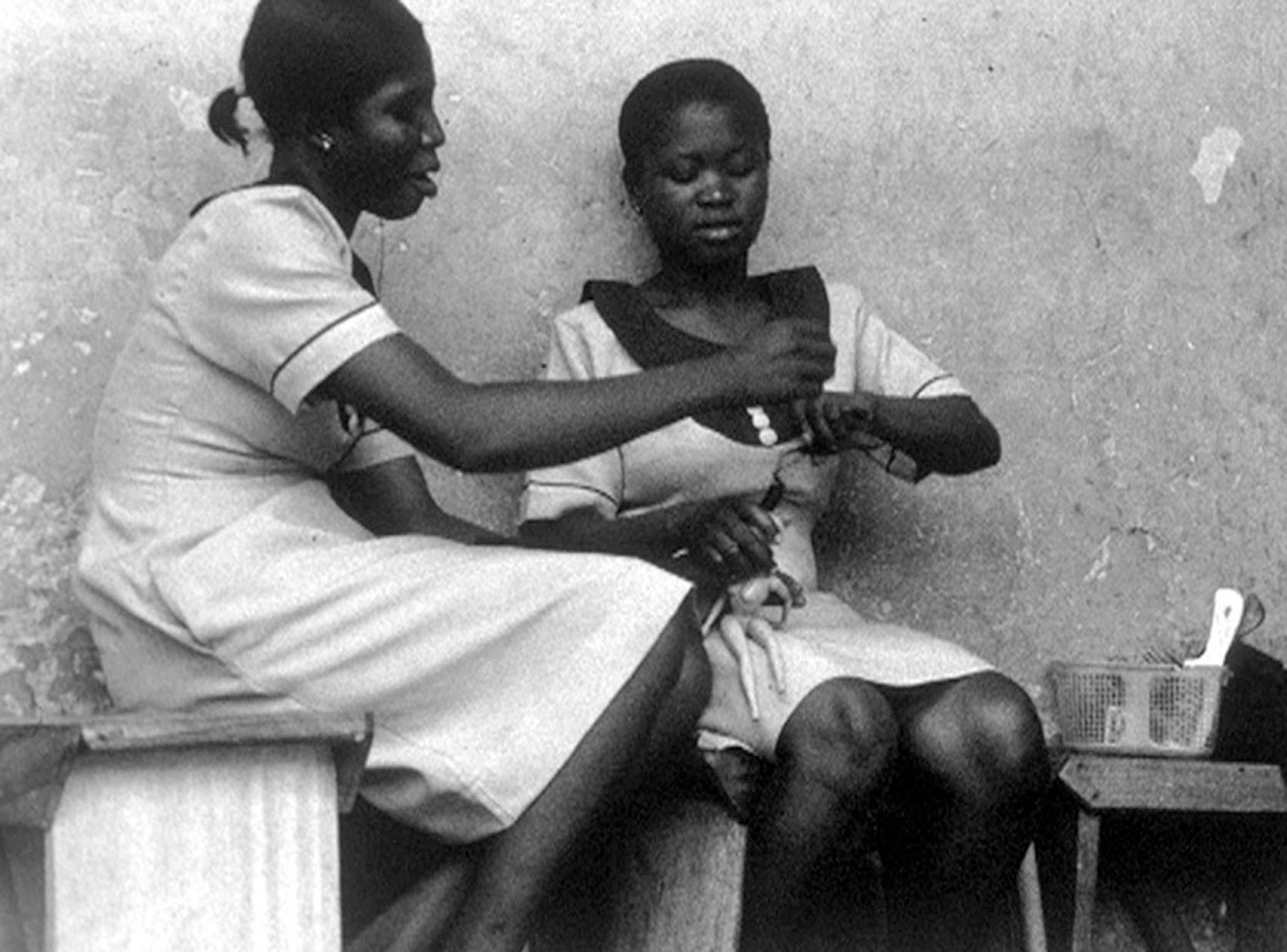 Two women sitting closely together, with one administering a vaccine to the other's upper arm. They are seated against a plain wall, and there is a small table with a basket containing medical supplies next to them. The image is black and white, suggesting it may be historical or taken in a setting with limited resources.