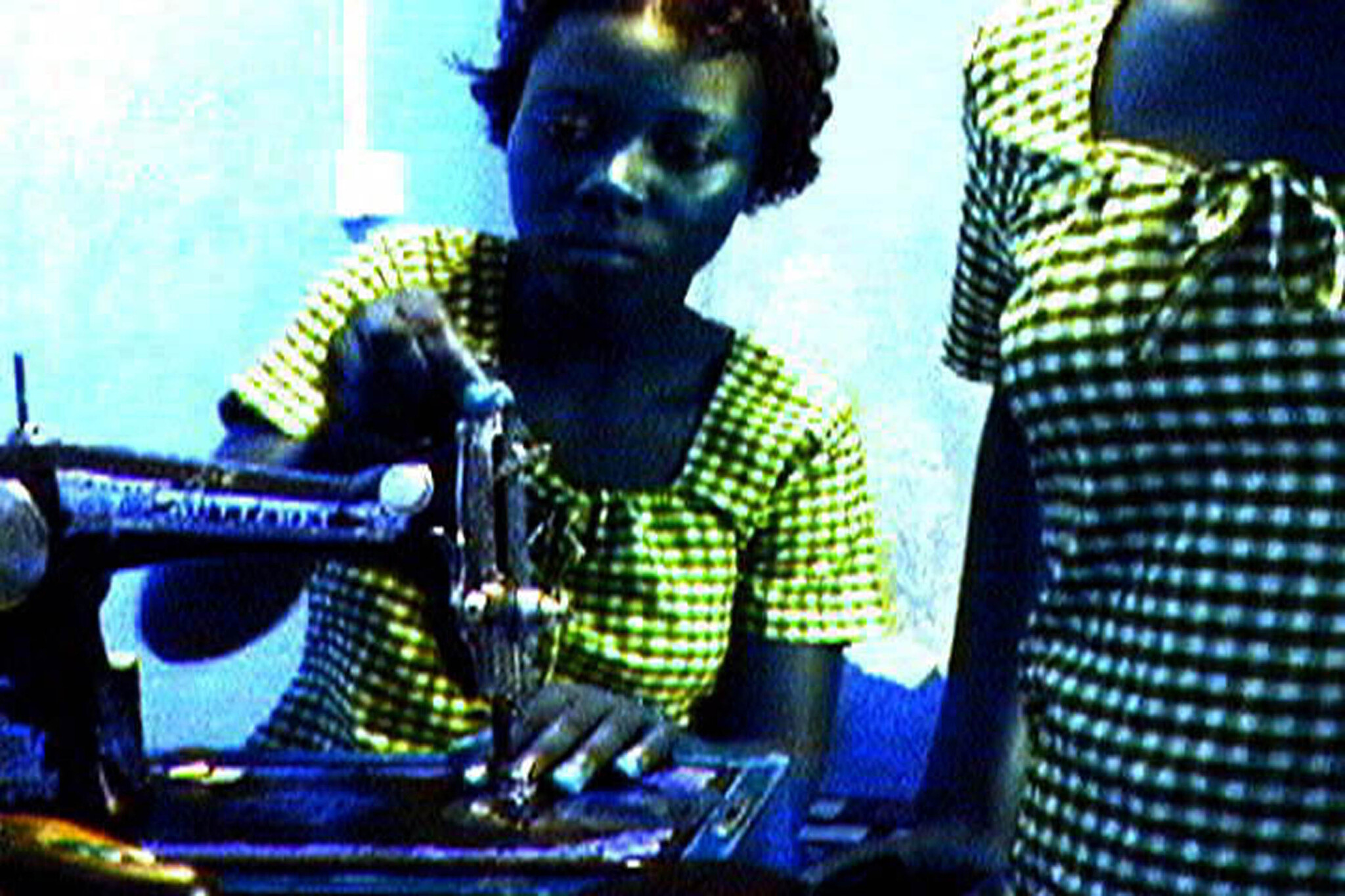 A person in a checkered shirt is operating an old-fashioned sewing machine with focus and attention to detail. The image has a blue tint and appears to be taken from a video with low resolution or from a screen, giving it a grainy texture. Another individual, partially visible on the right side, is wearing a similar patterned shirt.