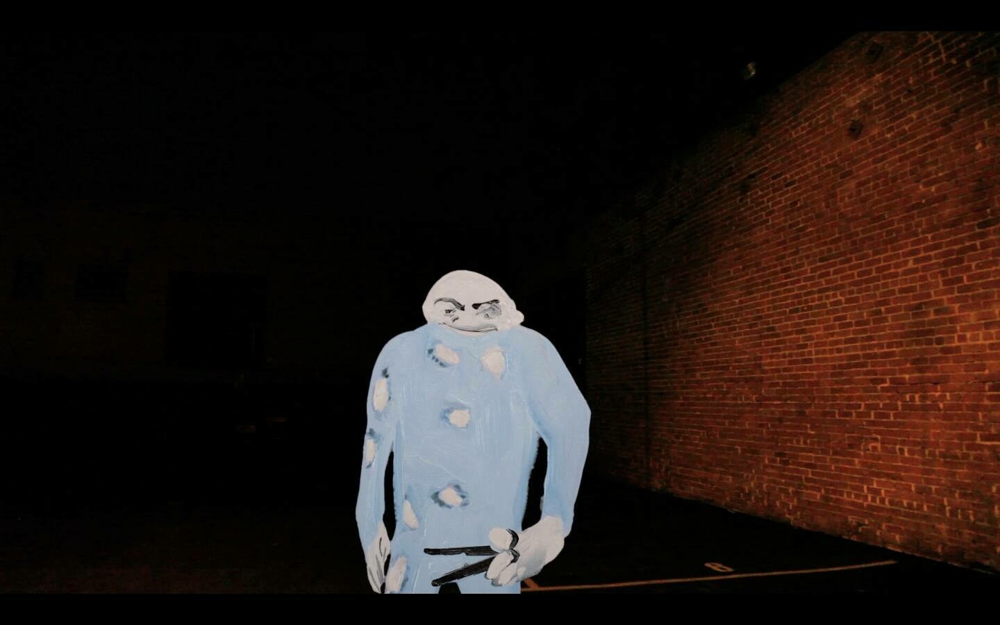 A person in a costume that resembles a cartoonish ghost with a grumpy expression stands in a dark alley with a brick wall in the background. The ghost costume is blue with multiple holes, and the person's face is visible through the head opening, painted to match the costume.