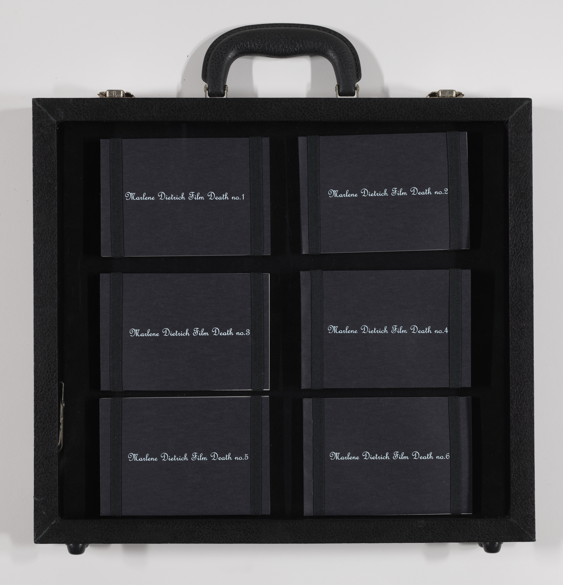 A black briefcase-style box with a handle on top, opened to reveal six compartments, each holding a black book with the title "Maxine Dietrich Film Death no.[number]" where [number] ranges from 1 to 6.
