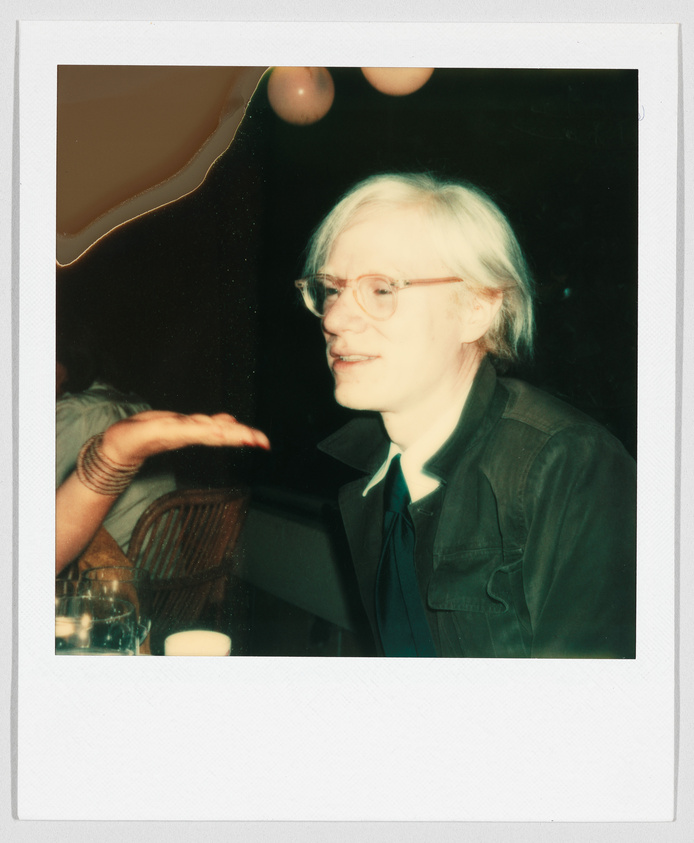 A person with light hair and glasses sits at a table, while another person gestures with their hand.