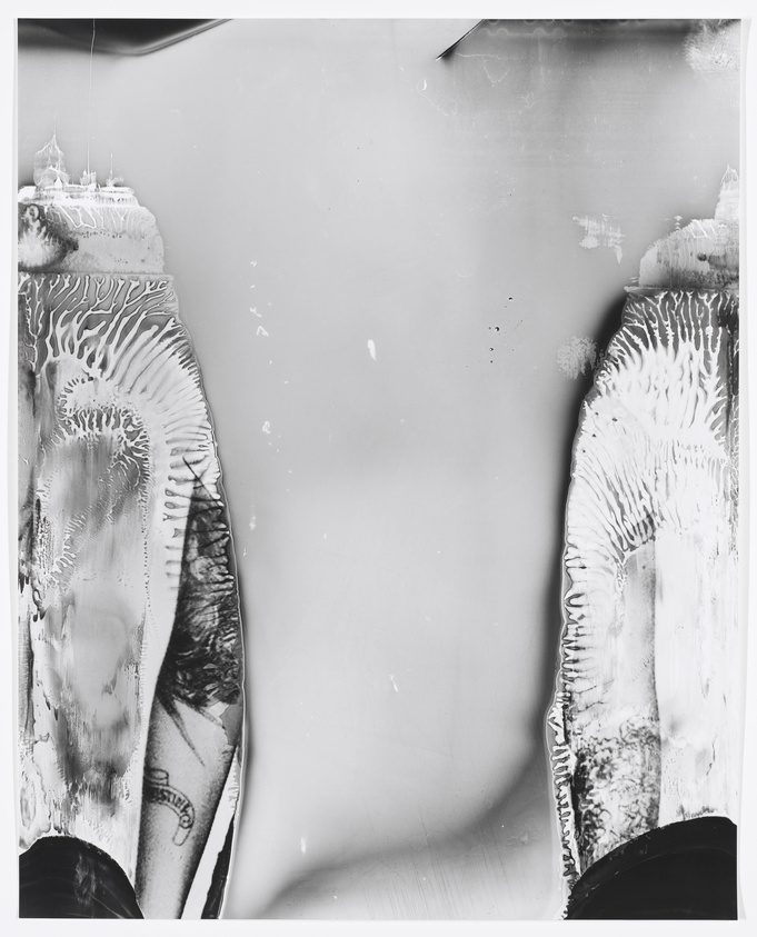 A black and white abstract image showing distorted and smeared photographic elements that resemble parts of human legs with tattoos, set against a blank background with some imperfections and creases.