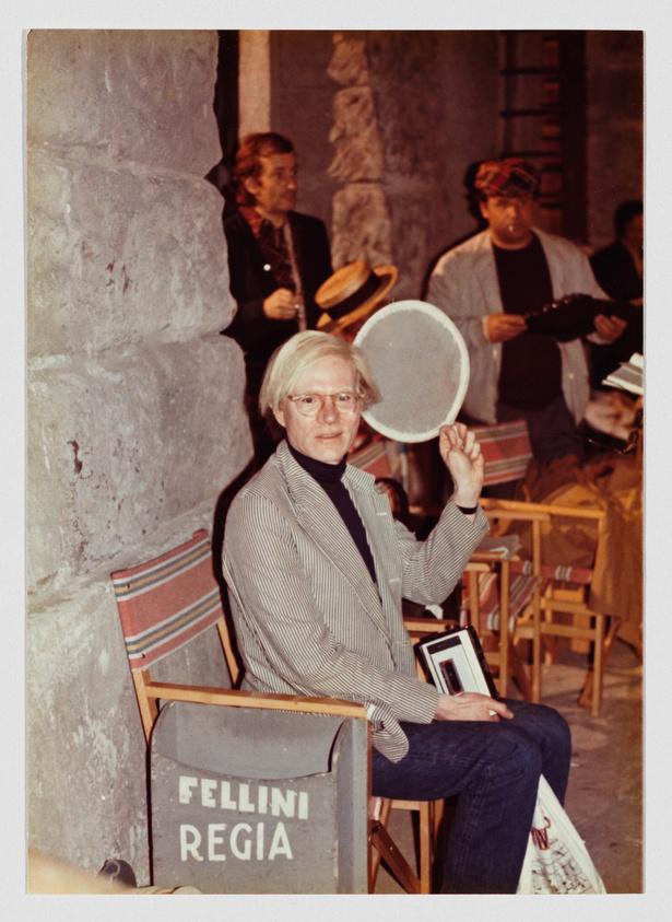 A man with light hair and glasses sits on a chair labeled "Fellini Regia," holding a cigarette.