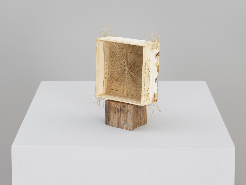 Sculpture of an open book with pages fanned out on a wooden pedestal, displayed on a white plinth.