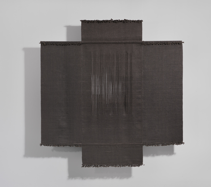 A textured black fabric artwork with frayed edges and a vertical white thread pattern on a gallery wall.