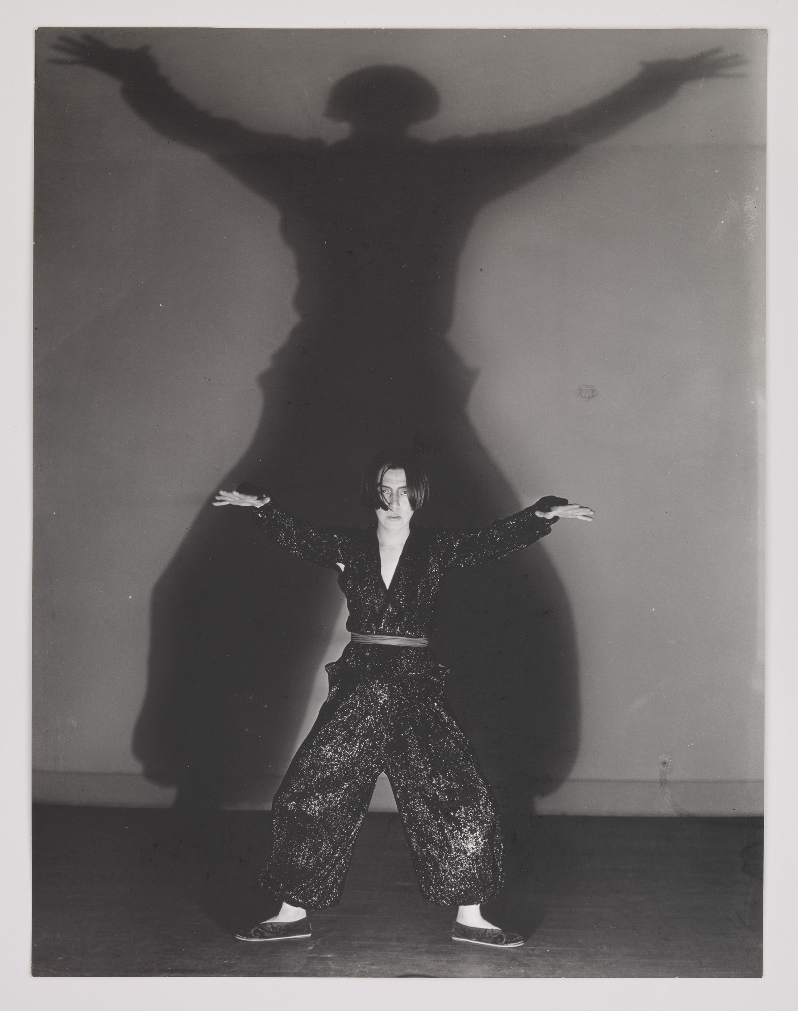 A black and white photograph of a person standing with arms outstretched, casting a large, dramatic shadow on the wall behind that mimics the pose. The person is wearing a patterned outfit and has a serious expression on their face.