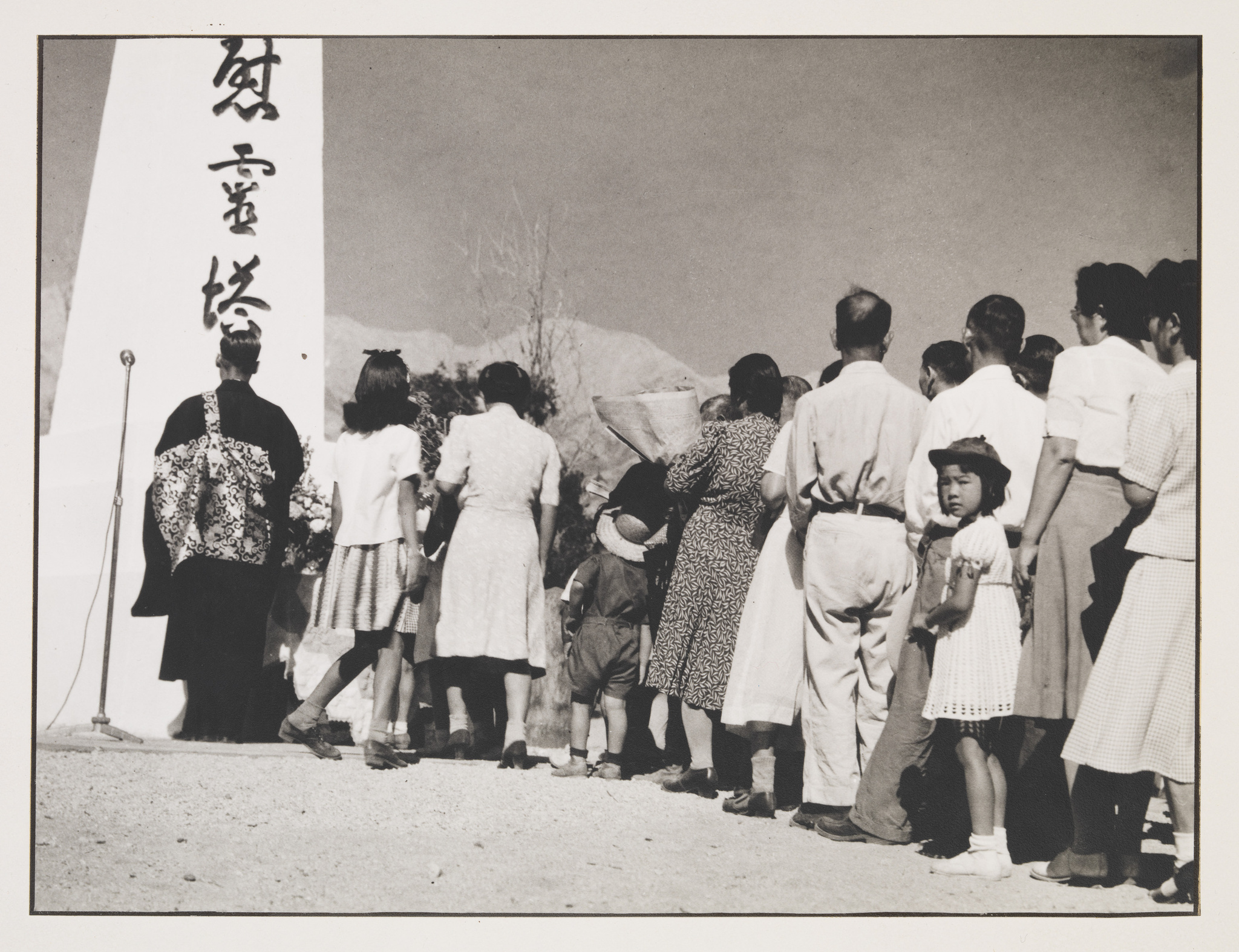 A black and white photograph of a group of people, including children and adults, gathered around a microphone stand next to a tall monument with Japanese characters. The people are dressed in a mix of Western and traditional Japanese attire, suggesting a cultural or commemorative event. A young girl in the foreground looks back towards the camera. The setting appears to be outdoors with a clear sky and sparse vegetation in the background.