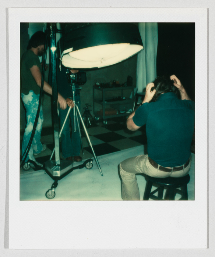 Two people in a photography studio, one adjusting a camera and the other sitting on a stool fixing hair.